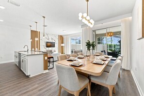 A modern kitchen with a central island, dining area with a wooden table and chairs, and a large window providing a view of the outdoors. - Chic Disney Escape, Themed Rooms & Private Pool (Four Corners)