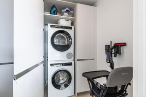 A compact laundry room with a front-loading washer and dryer, a small table, and a wall-mounted ironing board. - Mirador Estepona Hills 25 | Pool & Gym | Sea View (Estepona)