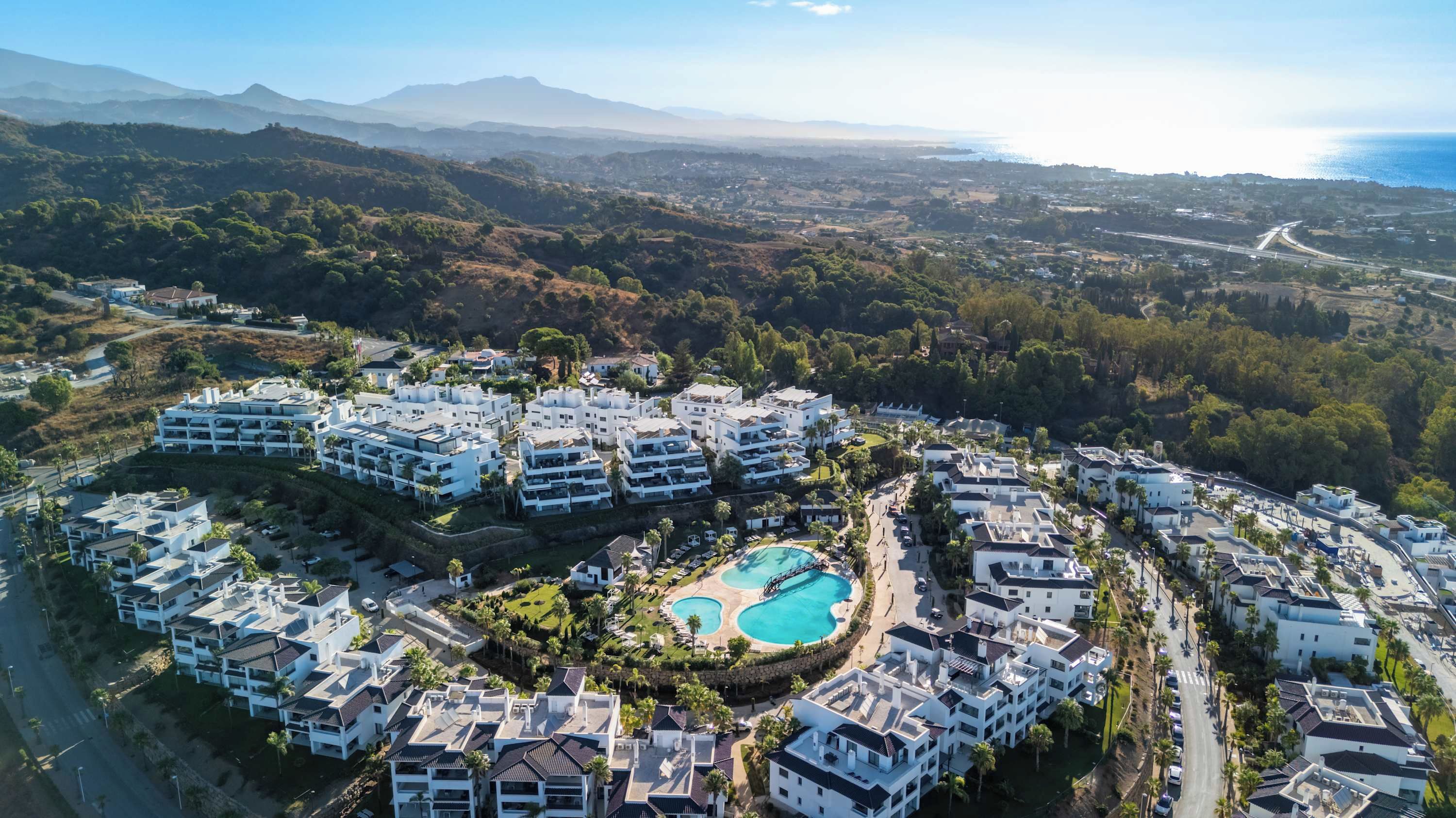 A resort with multiple white buildings, a central swimming pool, and a mountainous backdrop.