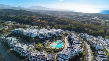 A resort with multiple white buildings, a central swimming pool, and a mountainous backdrop.