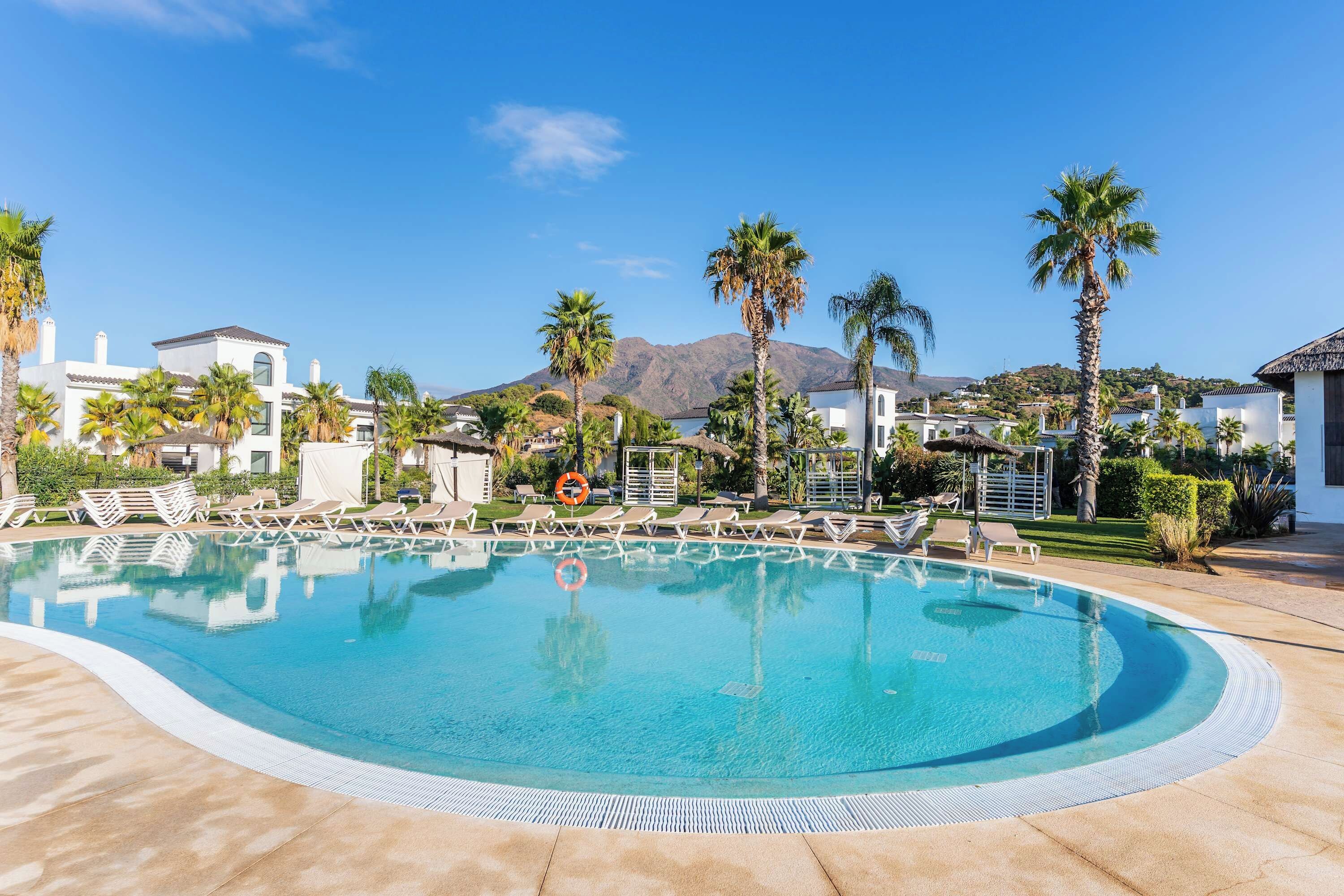 A clear blue swimming pool with lounge chairs and palm trees, surrounded by white buildings and a mountain in the background.