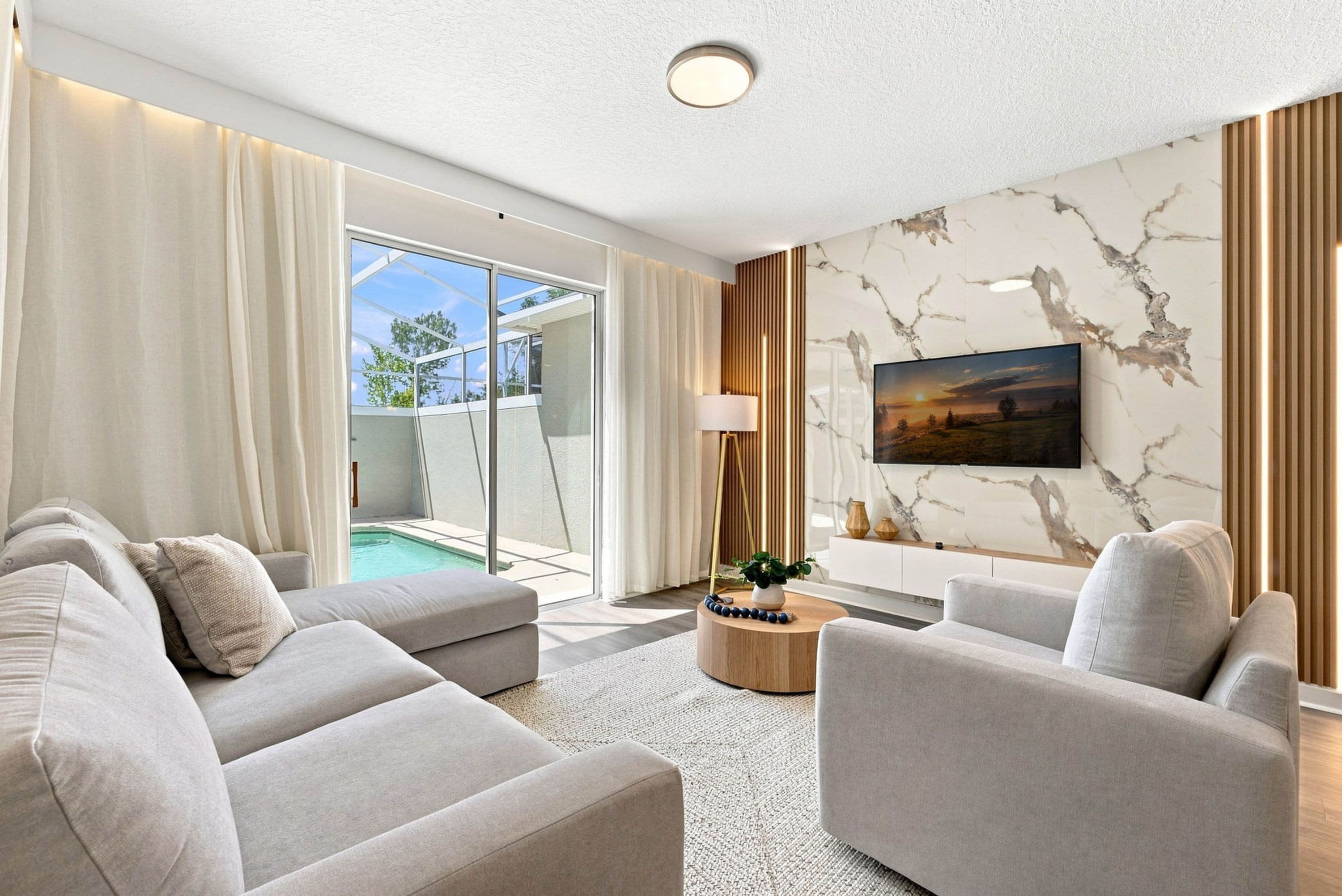 Un salon moderne comprenant un ensemble de canapés, une table basse, un téléviseur fixé au mur et donnant sur une piscine extérieure.