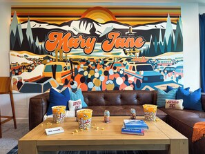 A cozy living room with a large mural, a brown leather couch, a wooden coffee table with popcorn and books, and a blue cushion. - Sleeps 18 | 2 Hot Tubs, Sauna & Game Room (Winter Park)