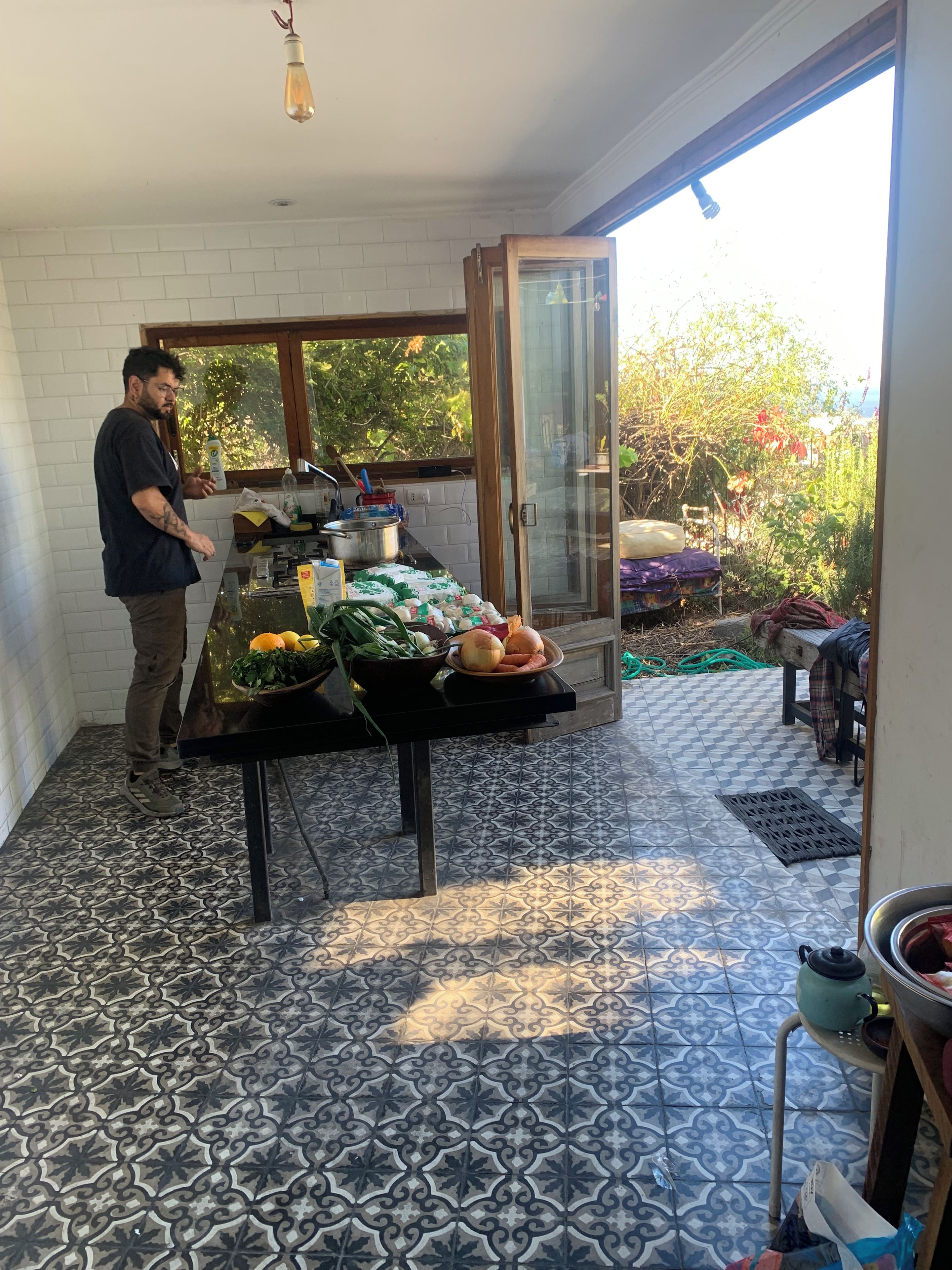 Une cuisine avec un sol carrelé, une table garnie de différents aliments et une fenêtre donnant sur un espace verdoyant.