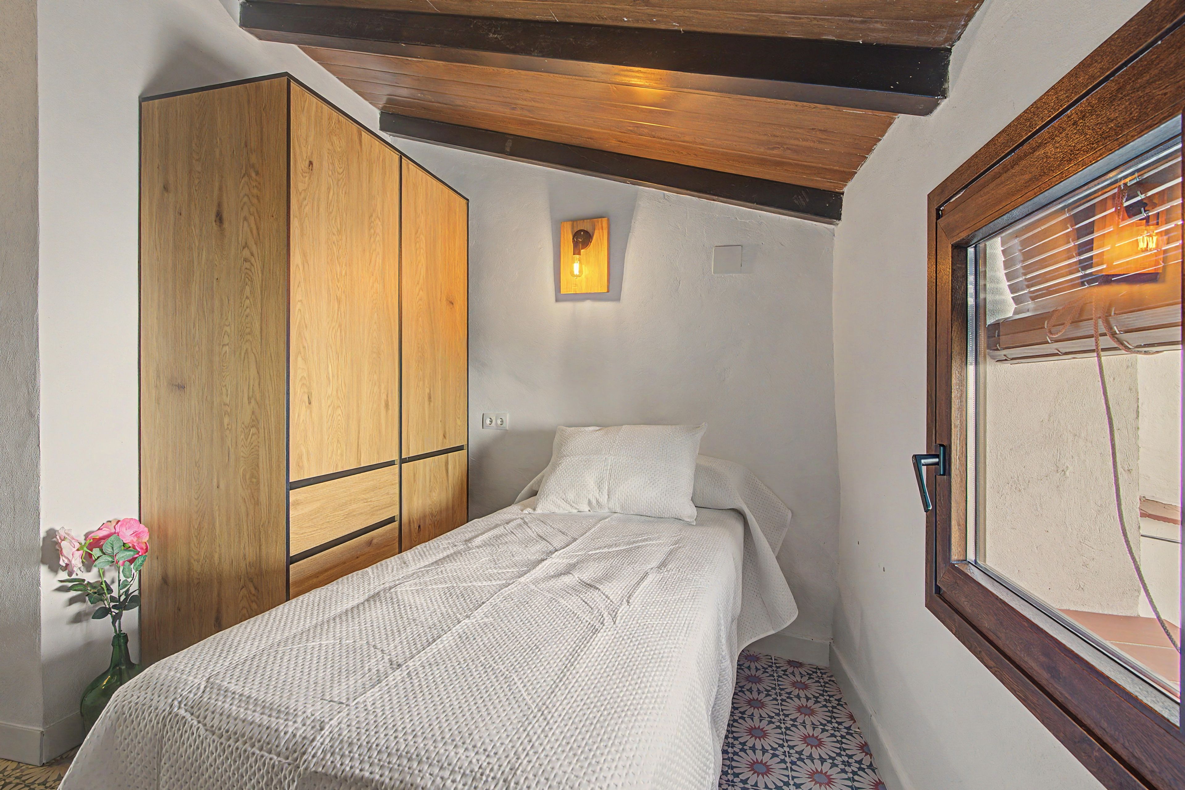 A small, single-bed room with a wooden wardrobe, a bedside table, and a window with blinds.