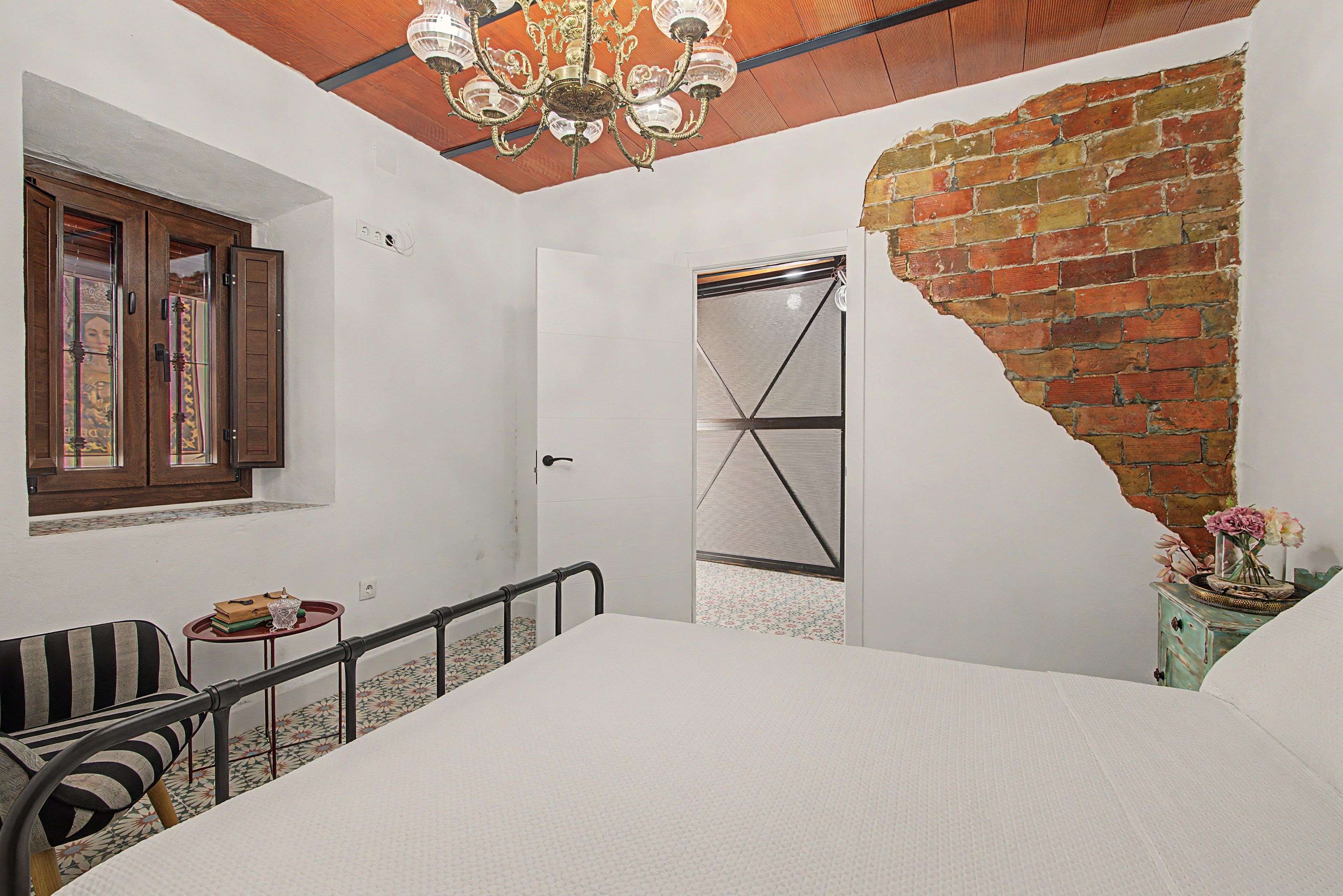 A room with a bed, a window with wooden shutters, a chandelier, a brick wall, and a small table with a vase.