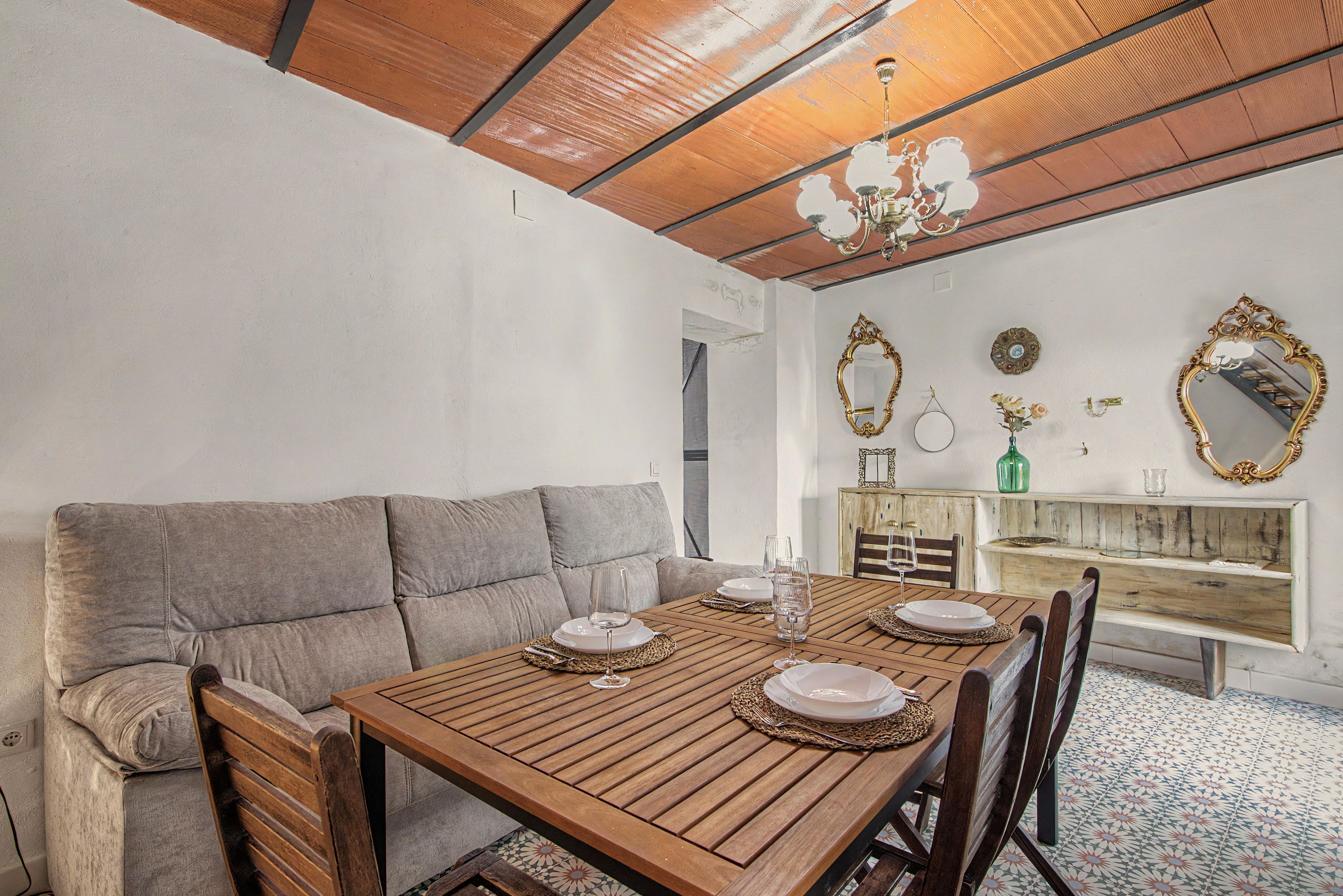 A dining area with a wooden table set for four, a grey sofa, and a wooden cabinet with decorative mirrors.