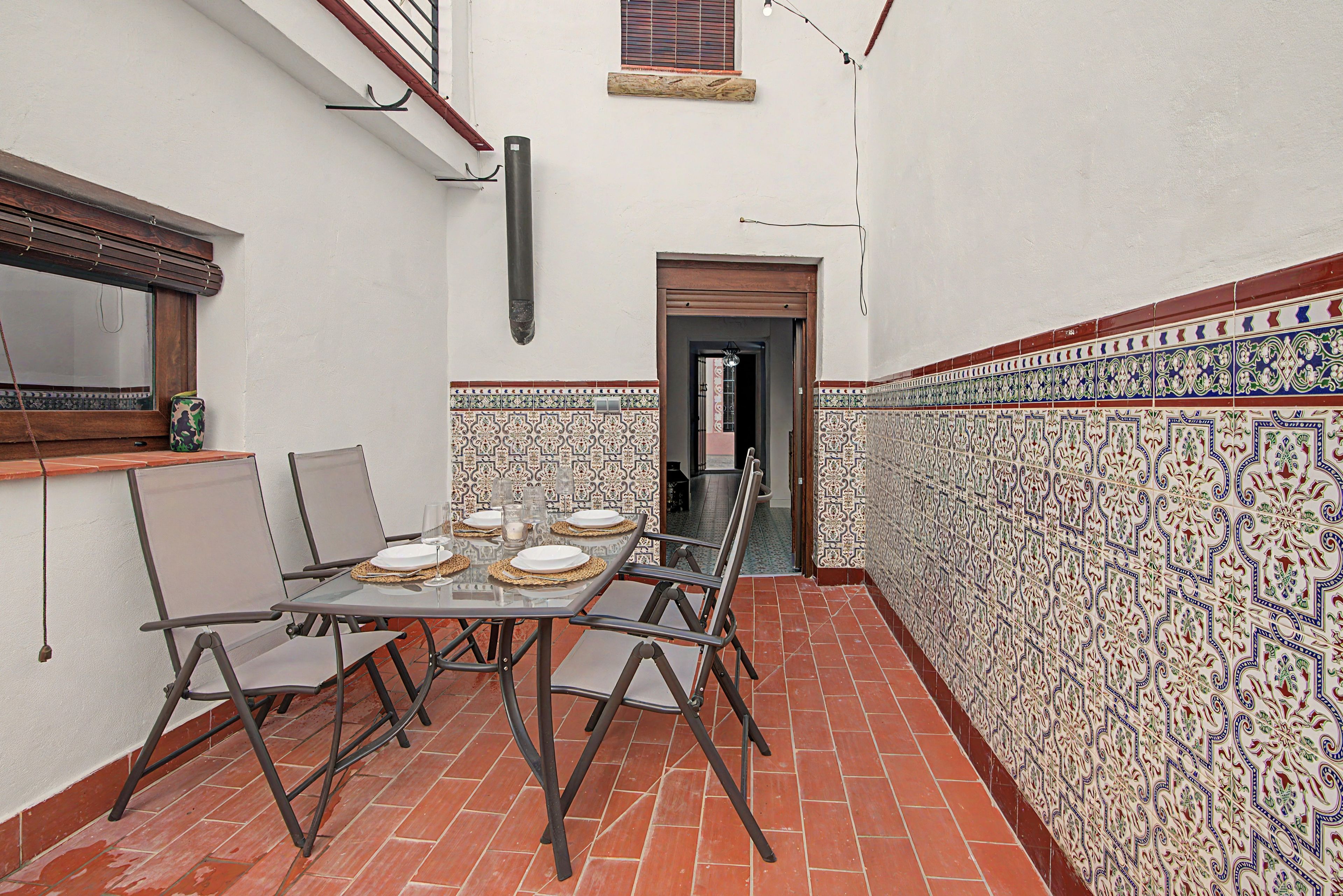 A narrow outdoor patio with a table set for four, surrounded by chairs, and a tiled wall with intricate patterns.