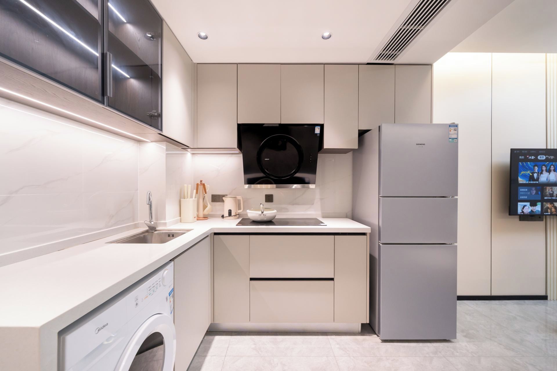 A modern kitchen with a built-in washing machine, a refrigerator, and a wall-mounted TV.