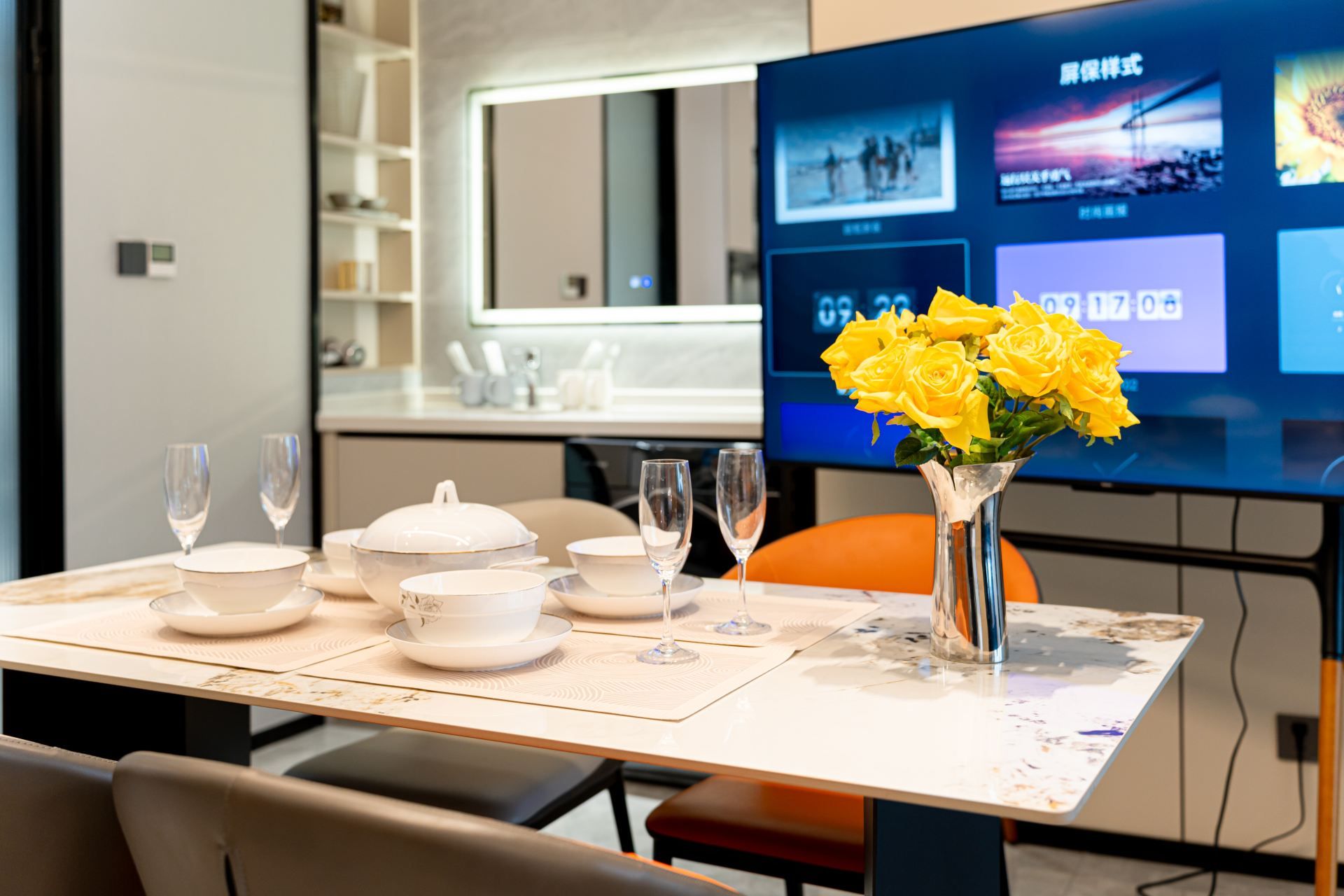 A dining table set with white dishes and glasses, a vase with yellow flowers, and a television displaying various images.