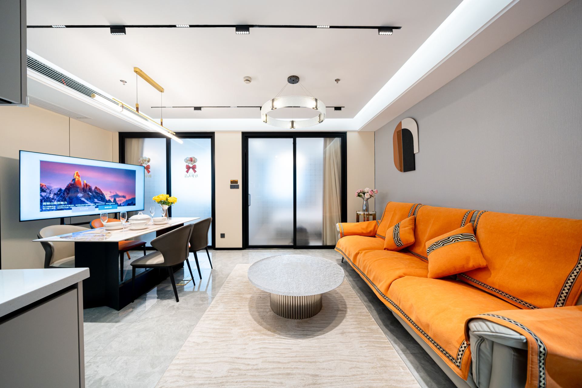 A modern living room with a flat-screen TV, a sofa, a coffee table, and a dining area with a table and chairs.
