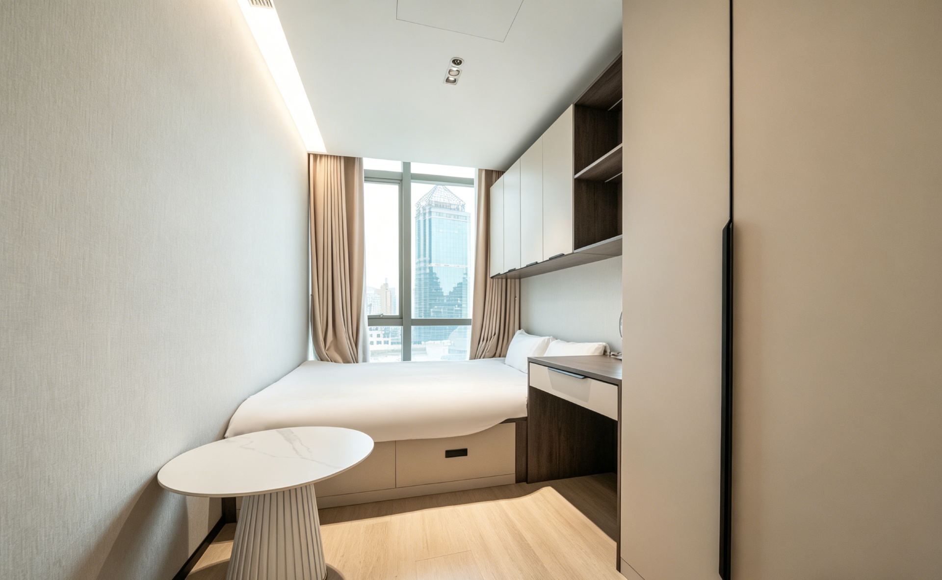 A compact hotel room with a bed, bedside table, and a view of the cityscape through large windows.