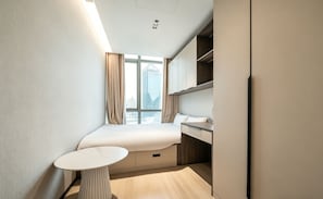A compact hotel room with a bed, bedside table, and a view of the cityscape through large windows.