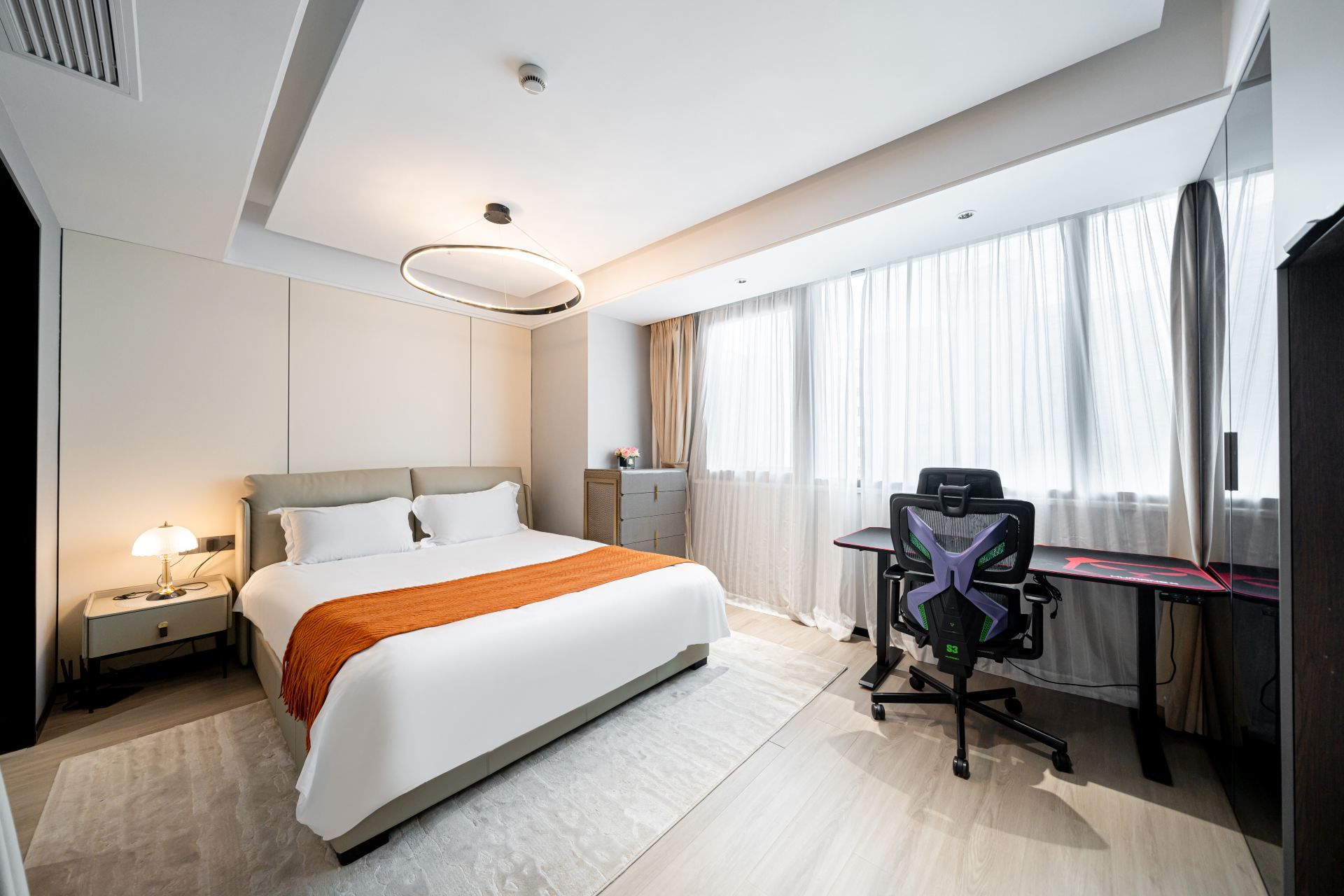 A hotel room with a bed, desk, chair, and a large window with curtains.