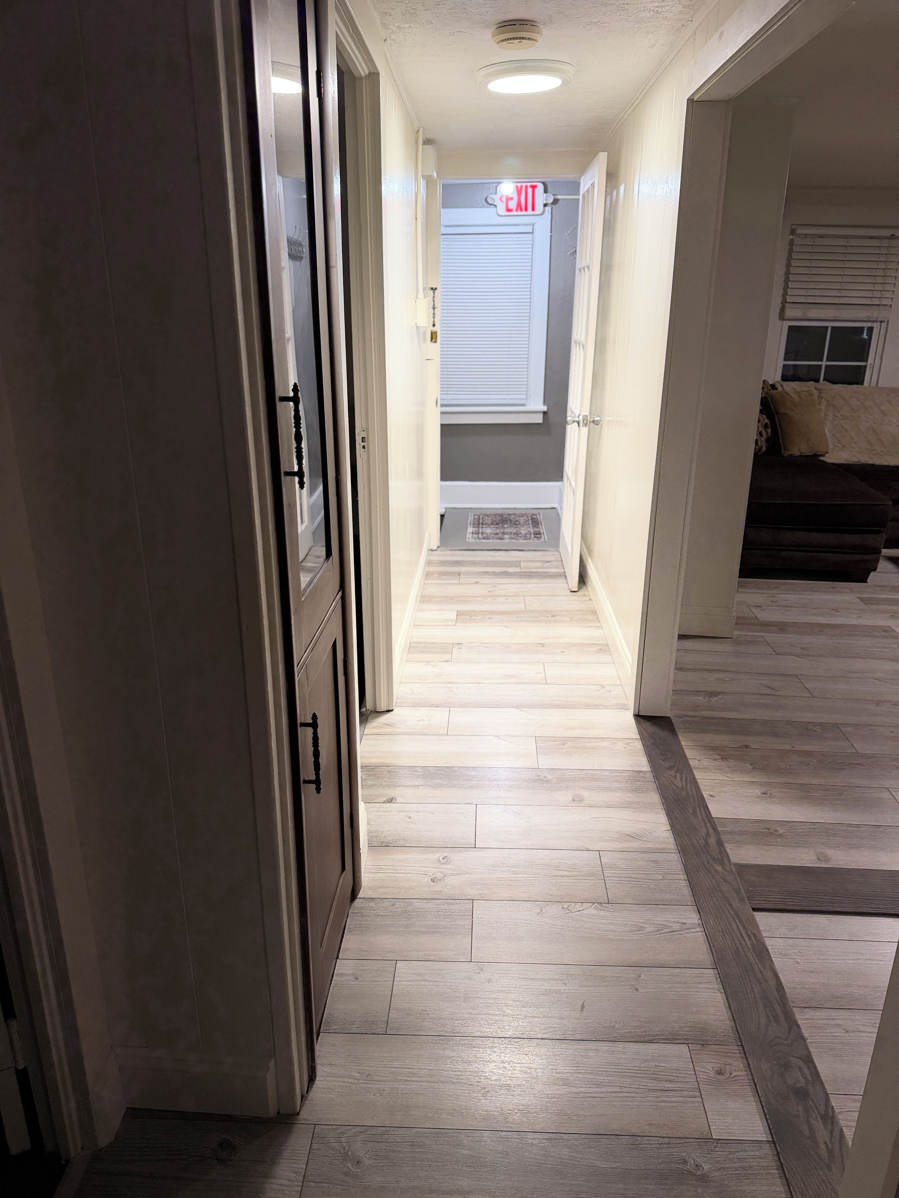 A hallway with a wooden floor, a door on the left, and an exit sign at the end.