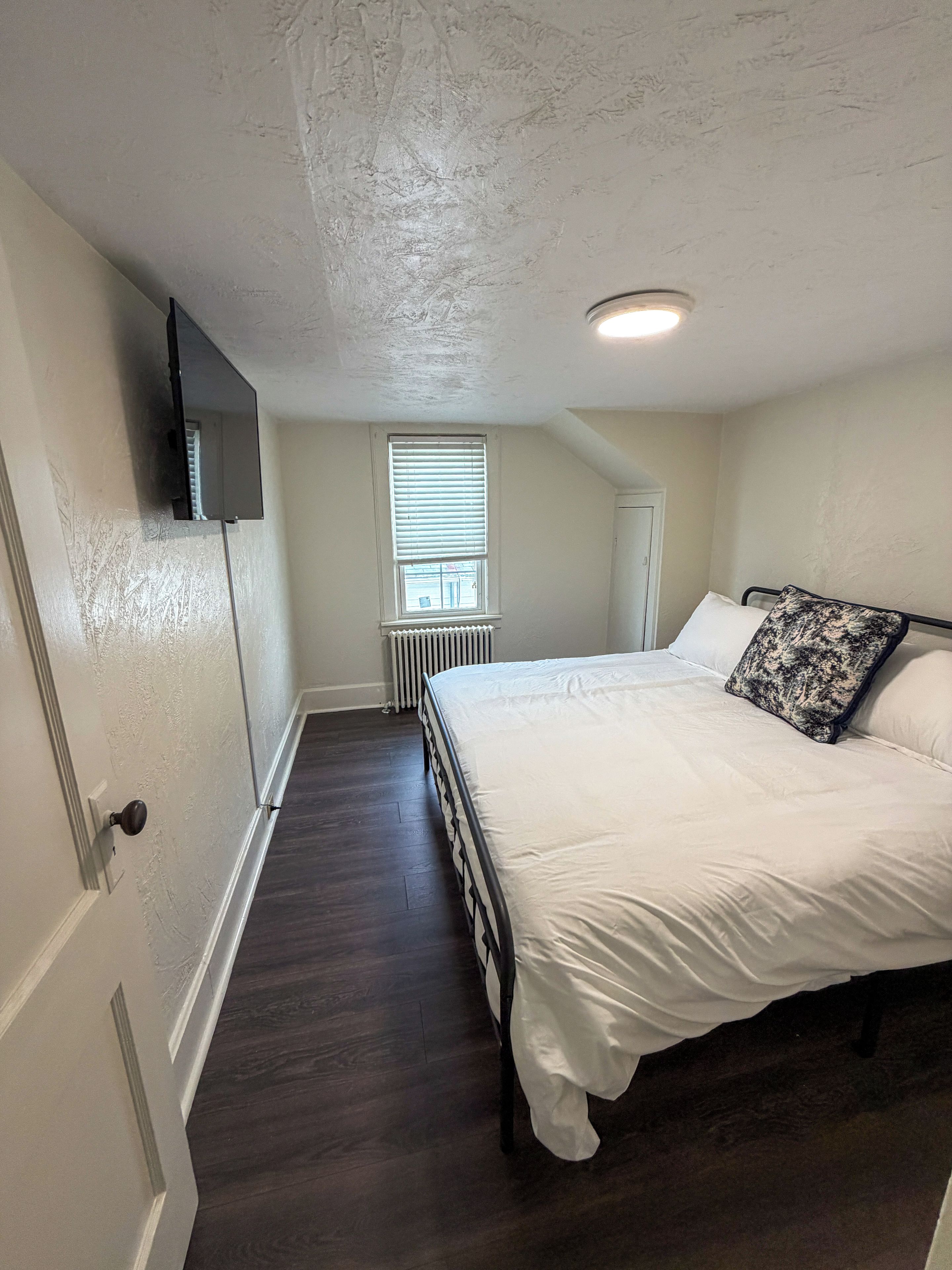 A bedroom with a bed, a television mounted on the wall, a window with blinds, and a radiator.