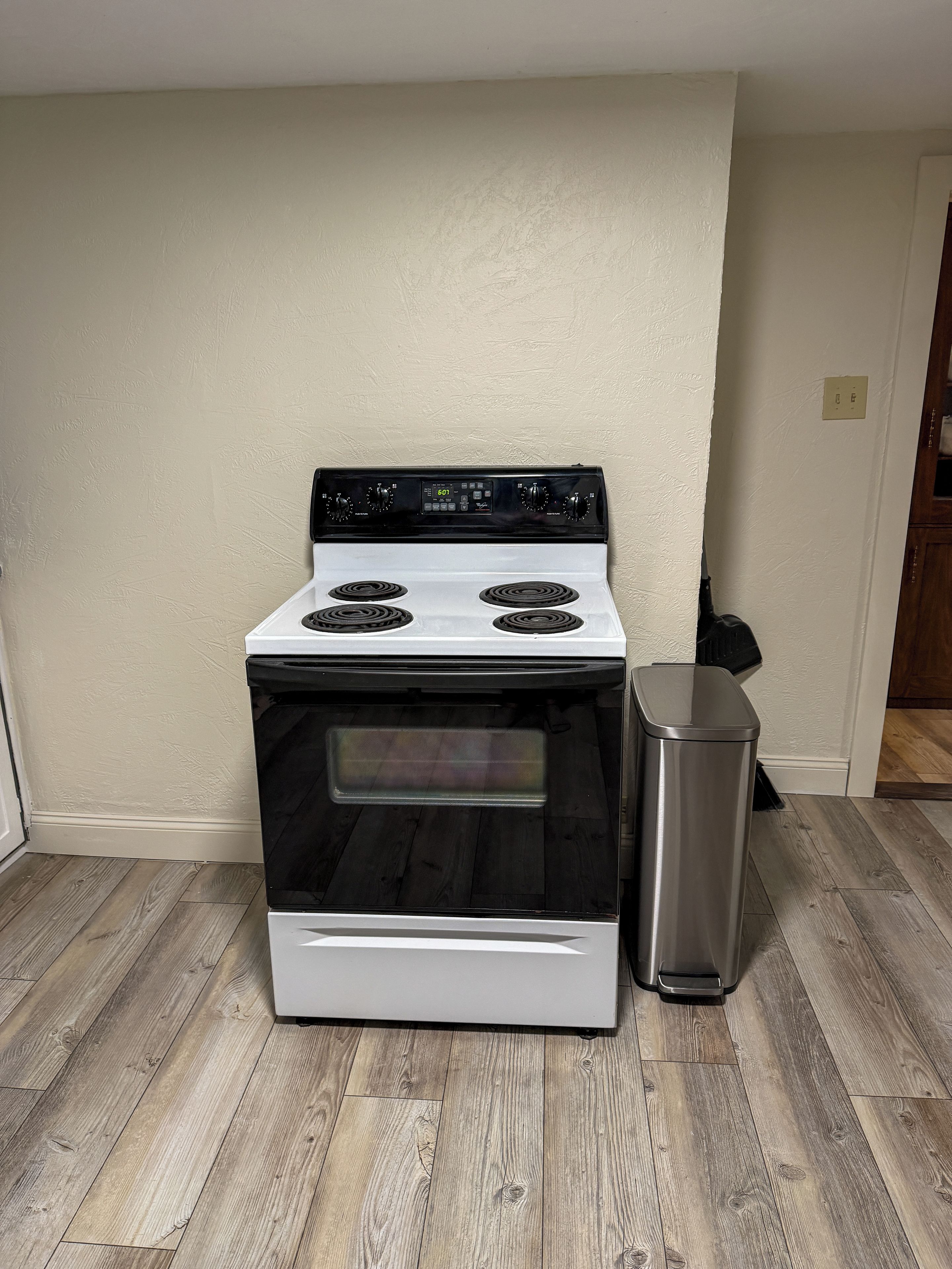 A kitchen with a white stove, a stainless steel trash can, and wooden flooring.