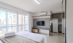 Ein modernes Hotelzimmer mit großem Fenster, Flachbildfernseher, Kühlschrank und einem Bett mit weißer Bettdecke.
