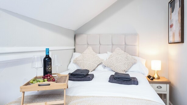 Ein Hotelzimmer mit einem Bett, Kissen, einem Nachttisch mit einer Flasche, Gläsern und einer Ablage mit Trauben.
