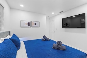 Ein Hotelzimmer mit einem Bett, einem Fenster mit Jalousien, einer Tür und zwei an der Wand befestigten Kunstwerken.