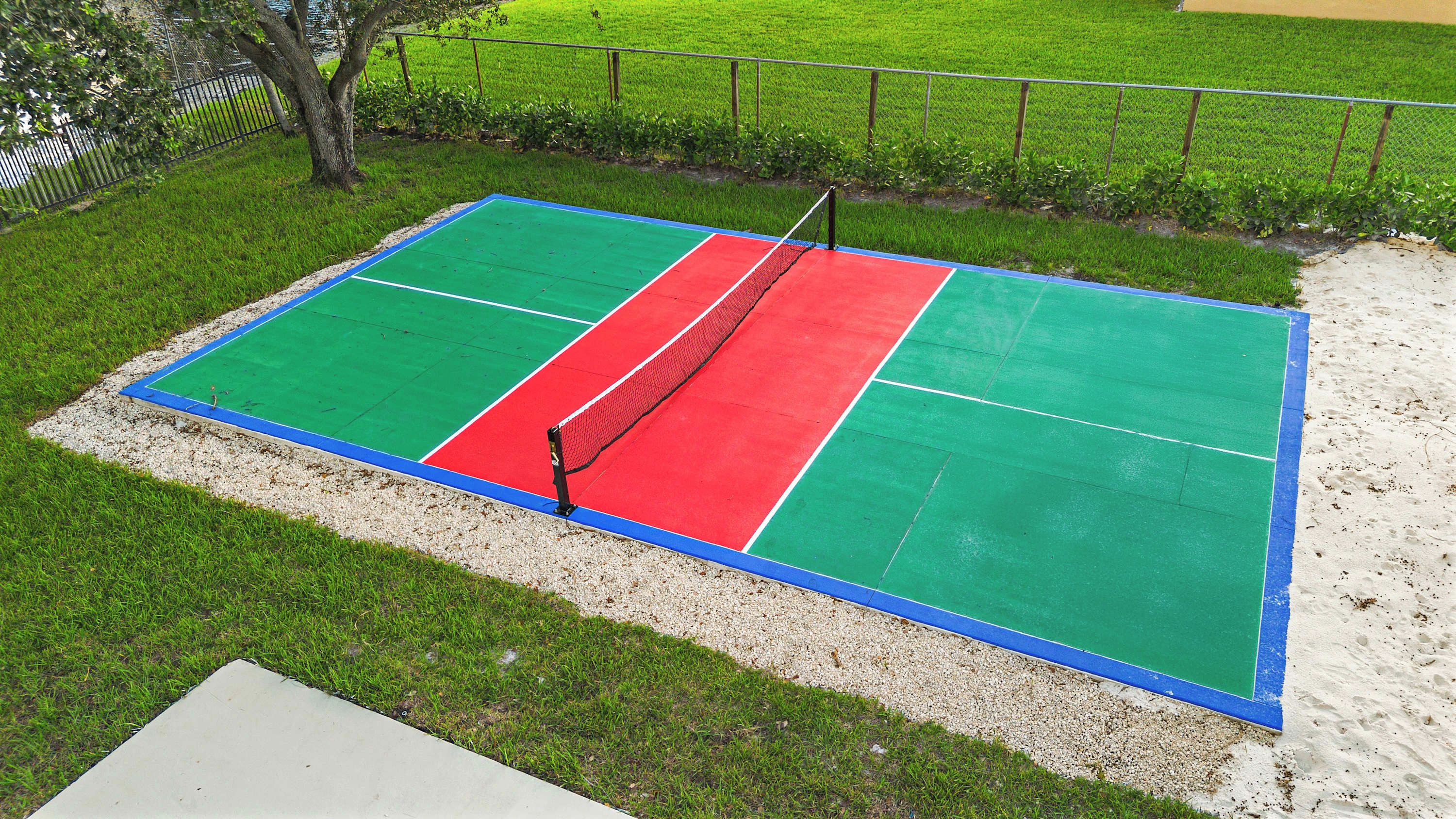 A tennis court with a net, surrounded by grass and a sandy area.