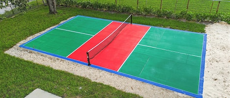 A tennis court with a net, surrounded by grass and a sandy area.