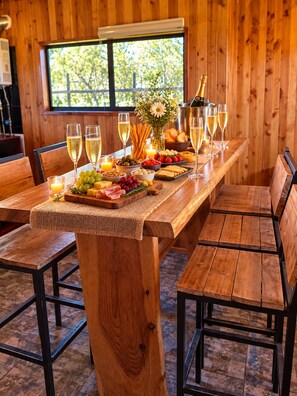 A wooden dining table set with glasses of champagne, a variety of cheeses, fruits, and bread, with a window in the background.