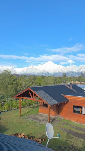 A wooden cabin with a metal roof, surrounded by a grassy area and trees, with snow-capped mountains in the background.