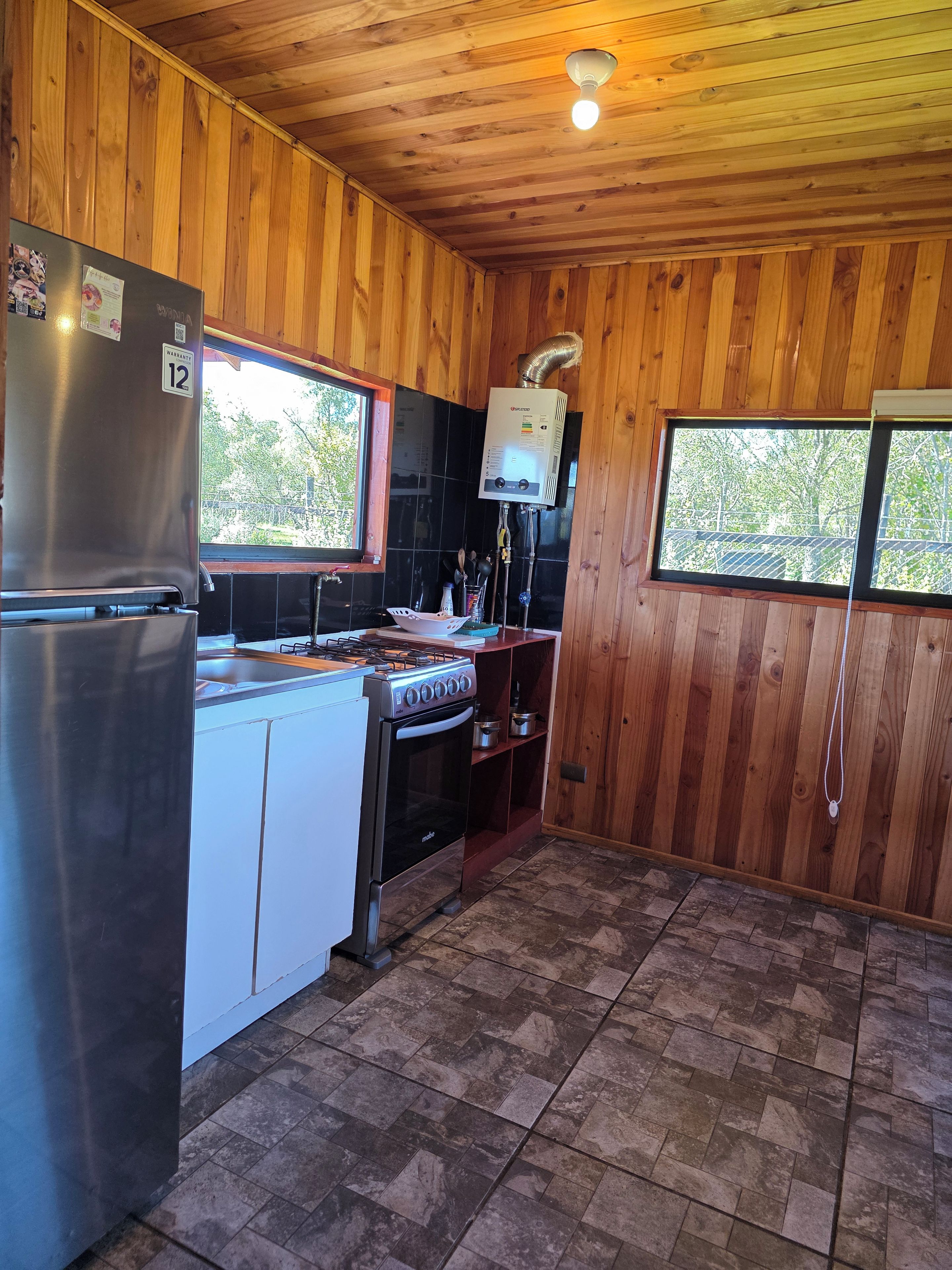 A compact kitchen with wooden walls, a stainless steel refrigerator, a stove, and a built-in oven.