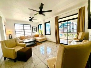 Ein geräumiges Wohnzimmer mit einer Sofagarnitur, zwei Sesseln, einem Couchtisch und einem Deckenventilator. Große Fenster sorgen für natürliches Licht und bieten einen Ausblick nach draußen.