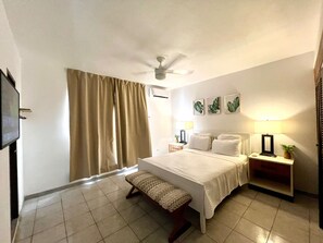 Habitación de hotel con cama, una banca, ventilador de techo, mesita de noche con lámpara y televisor montado en la pared.