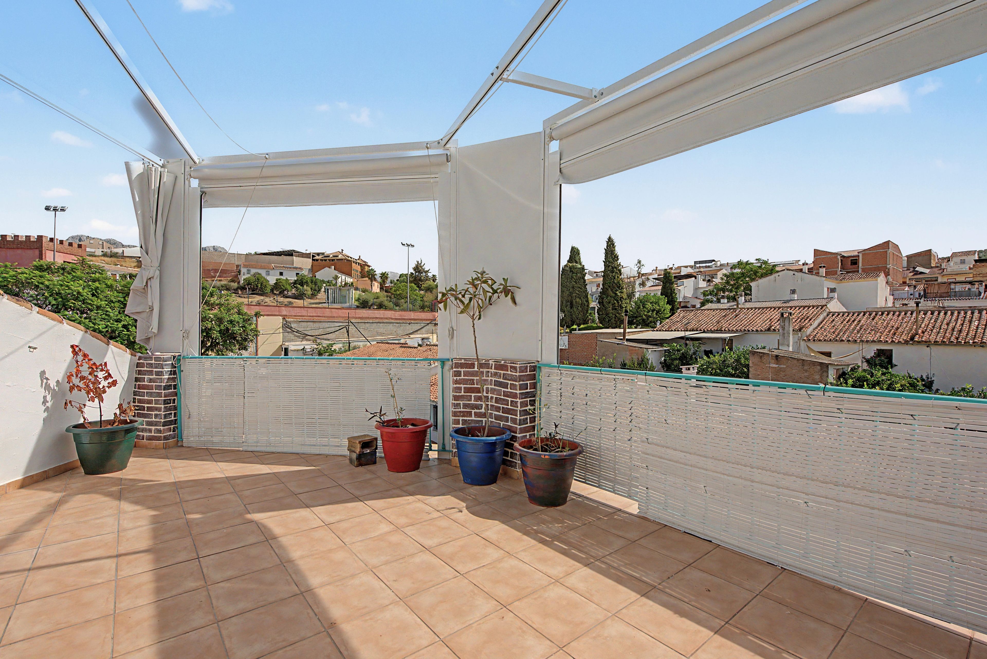 A covered balcony with tiled flooring, potted plants, and a view of a residential area with buildings and trees.