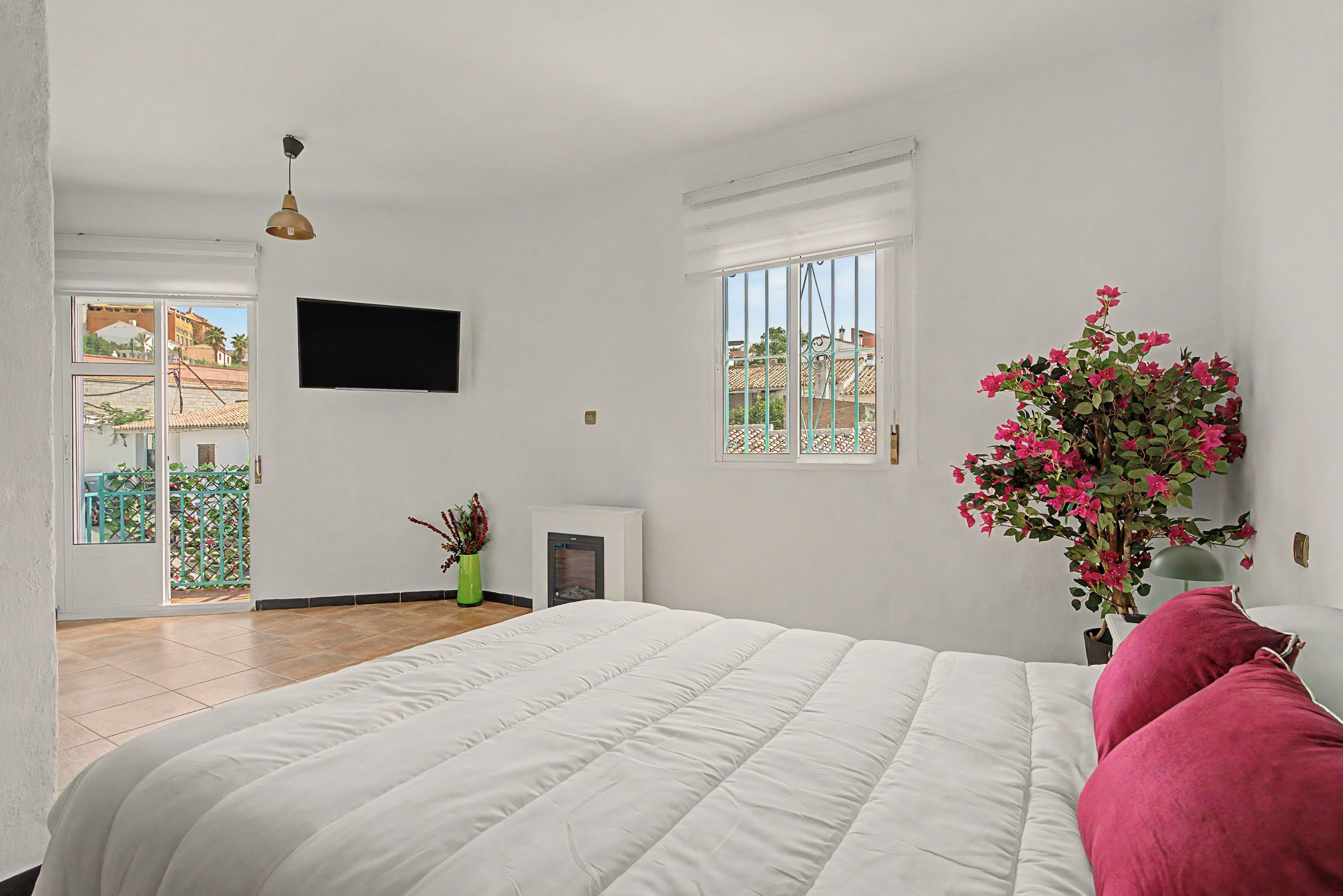 Ein Schlafzimmer mit einem Bett, einem an der Wand befestigten Fernseher, einem Fenster mit Blick auf Gebäude und einer Topfpflanze mit rosa Blüten.
