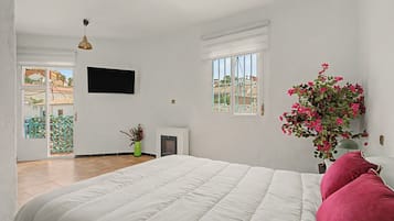 Ein Schlafzimmer mit einem Bett, einem an der Wand befestigten Fernseher, einem Fenster mit Blick auf Gebäude und einer Topfpflanze mit rosa Blüten.