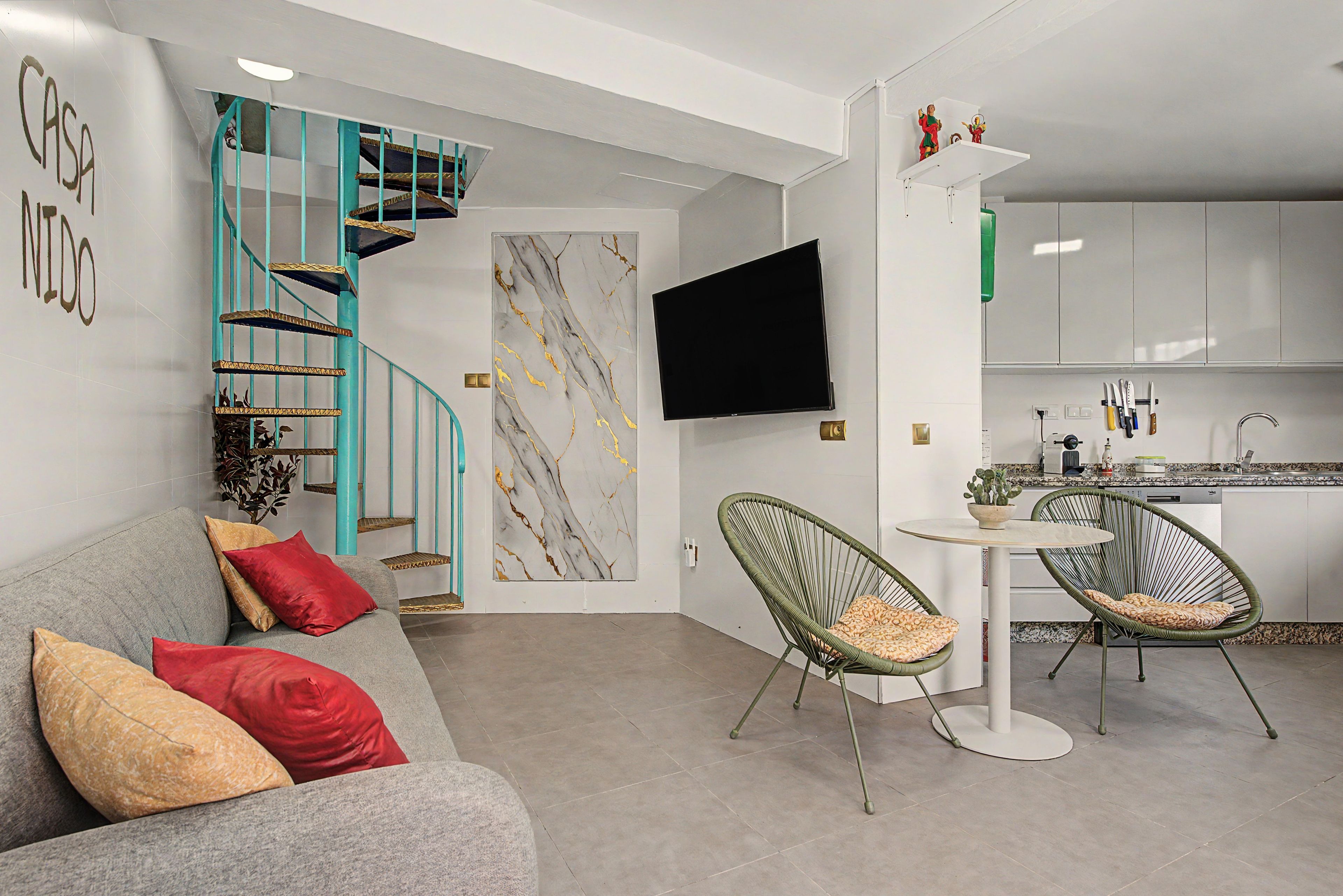 Ein moderner Wohnraum mit grauem Sofa, roten Kissen, einer Marmorwand, einem Flachbildfernseher, einer Küchenzeile mit weißen Schränken und einer türkisfarbenen Wendeltreppe.