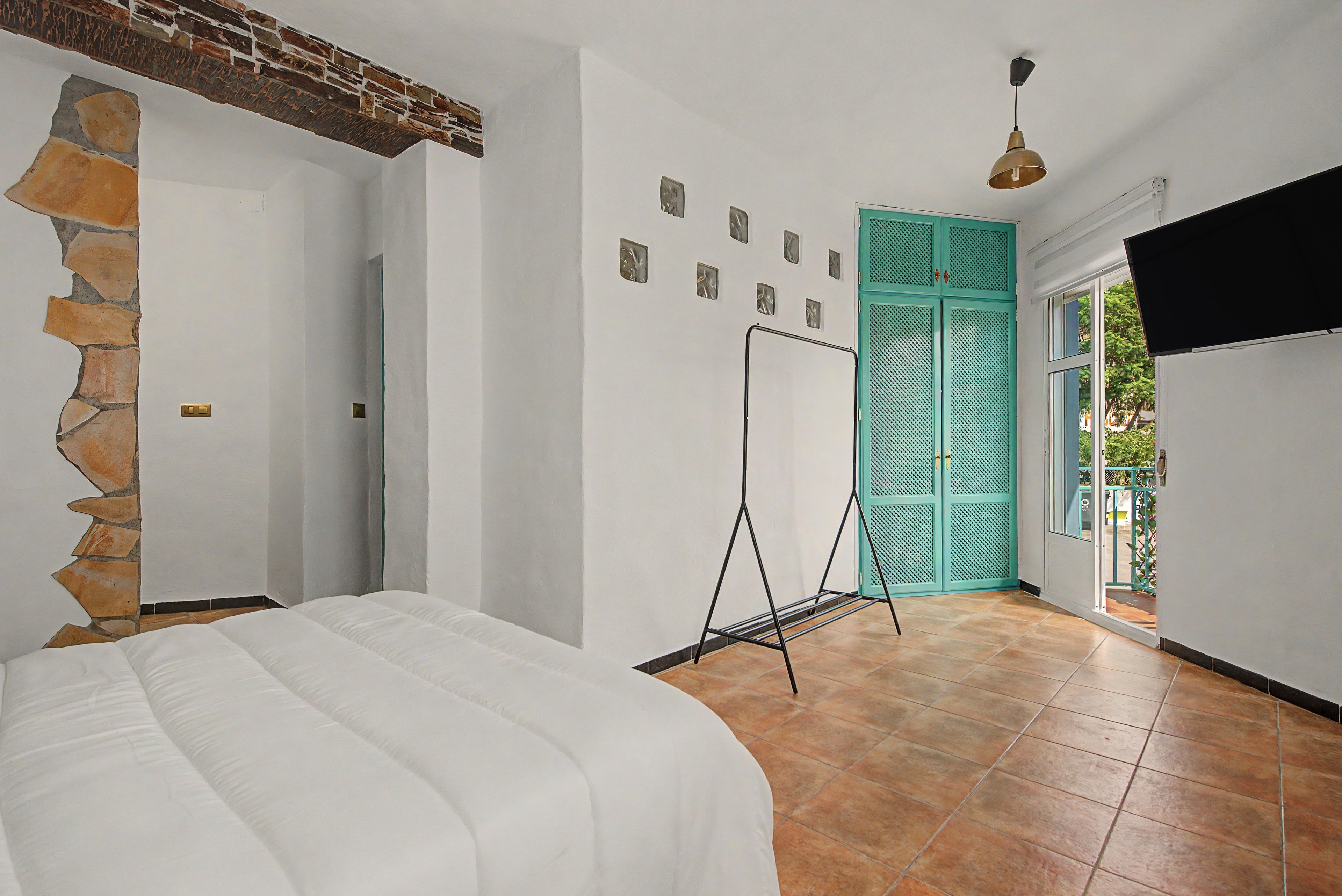 Una habitación con una cama, una pared de piedra, una puerta verde azulado y un televisor sobre un soporte.