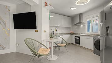 Ett modernt kök med vita skåp, en inbyggd ugn, en diskmaskin och ett kylskåp. Det finns en matplats med ett runt bord och två fåtöljer i rotting.