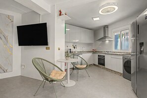 Una cucina moderna con mobili bianchi, forno integrato, lavastoviglie e frigorifero. C'è un'area pranzo con un tavolo rotondo e due sedie in vimini.