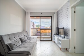 A modern living room with a grey sofa, a TV on a white stand, and a balcony with a view of buildings. - Apartment w/ Barbecue in Building w/ Pool Roh0304 (Florianópolis)