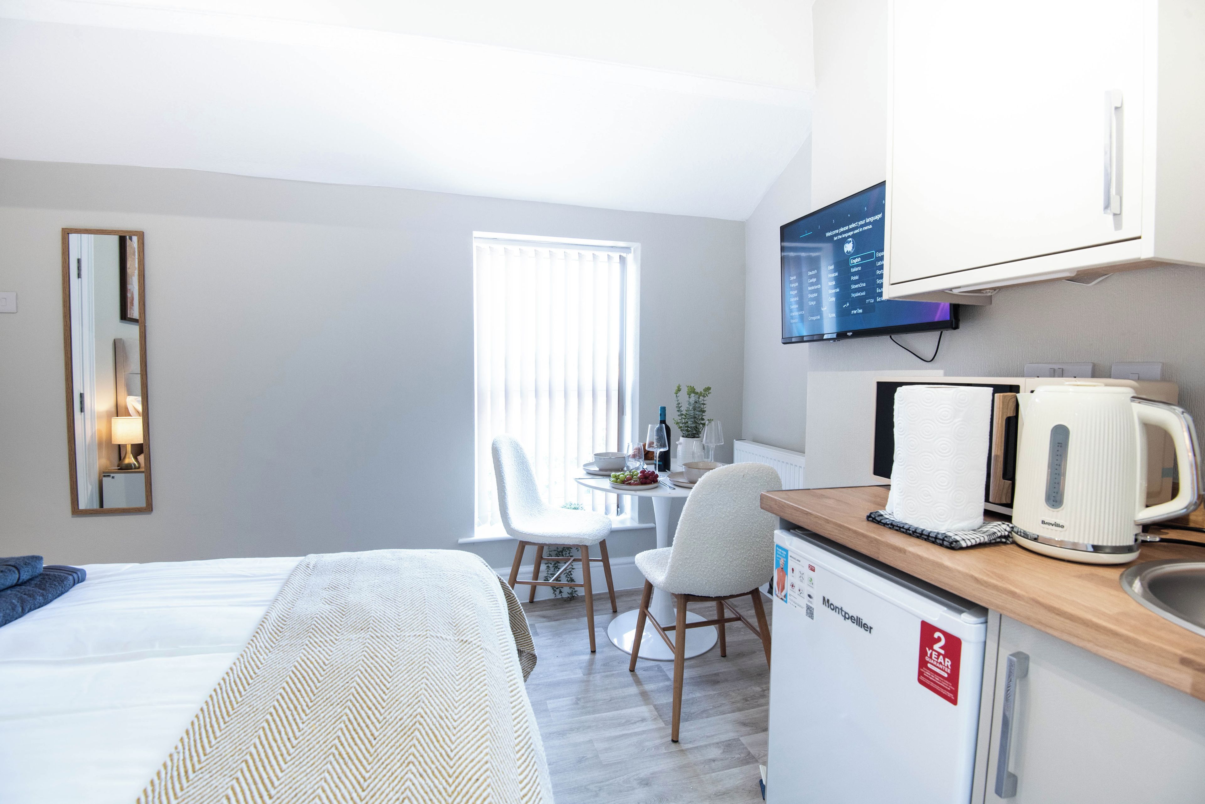 A hotel room with a bed, a desk with a kettle, a television, and a window with blinds.