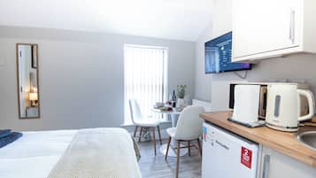 A hotel room with a bed, a desk with a kettle, a television, and a window with blinds.