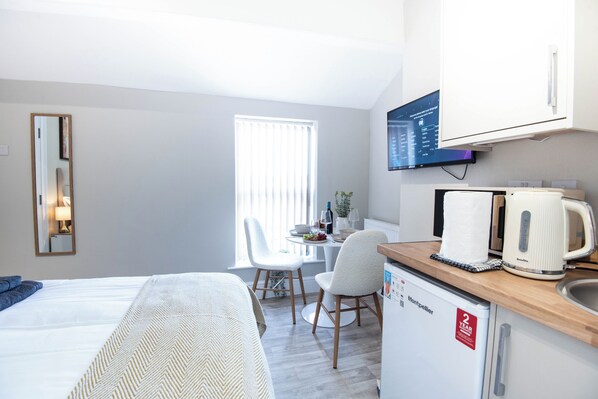 A hotel room with a bed, a desk with a kettle, a television, and a window with blinds.