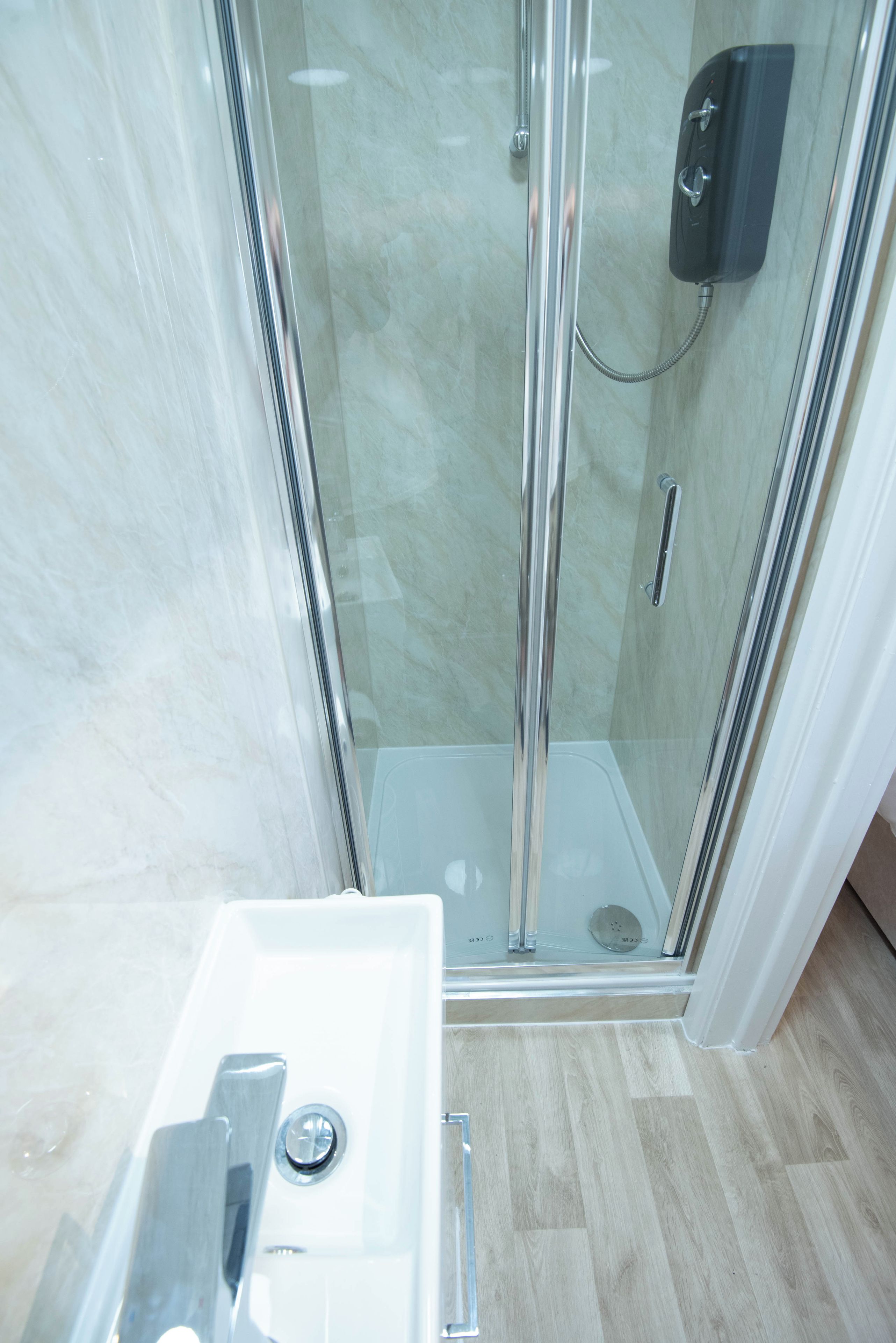 A glass-enclosed shower with a handheld showerhead, a white sink with a chrome faucet, and a wooden floor.