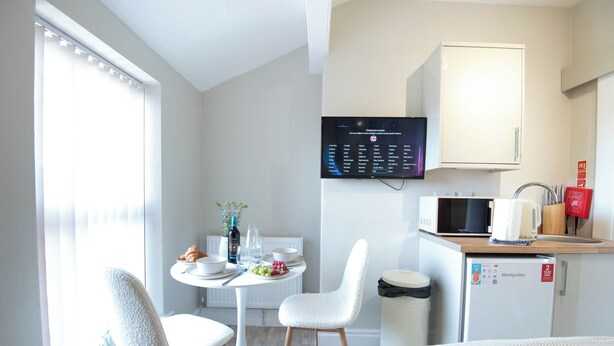 Ein kompaktes Hotelzimmer mit einer Küchenzeile, einem Essbereich mit Tisch und Stühlen, einem flachbildfernseher an der Wand und einem Fenster mit Jalousien.