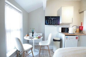 A compact hotel room with a kitchenette, a dining area with a table and chairs, a flat-screen TV mounted on the wall, and a window with blinds.
