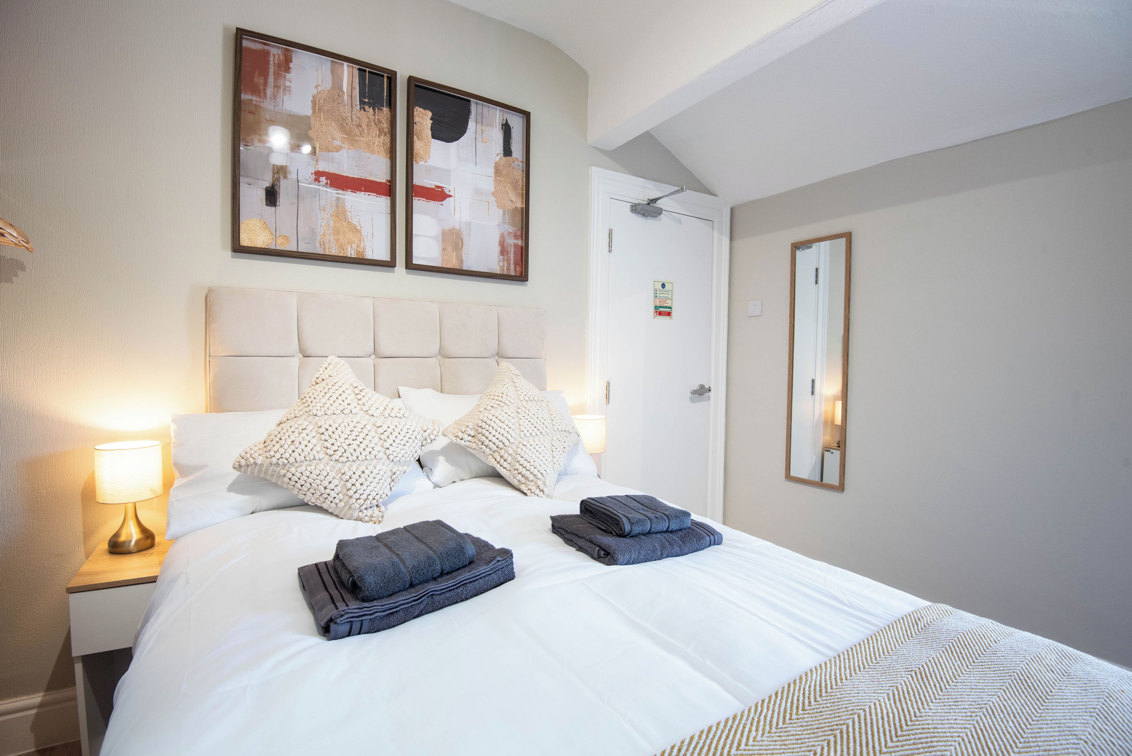 A hotel room with a bed, two pillows, a bedside lamp, folded towels, a door, and two framed pictures on the wall.