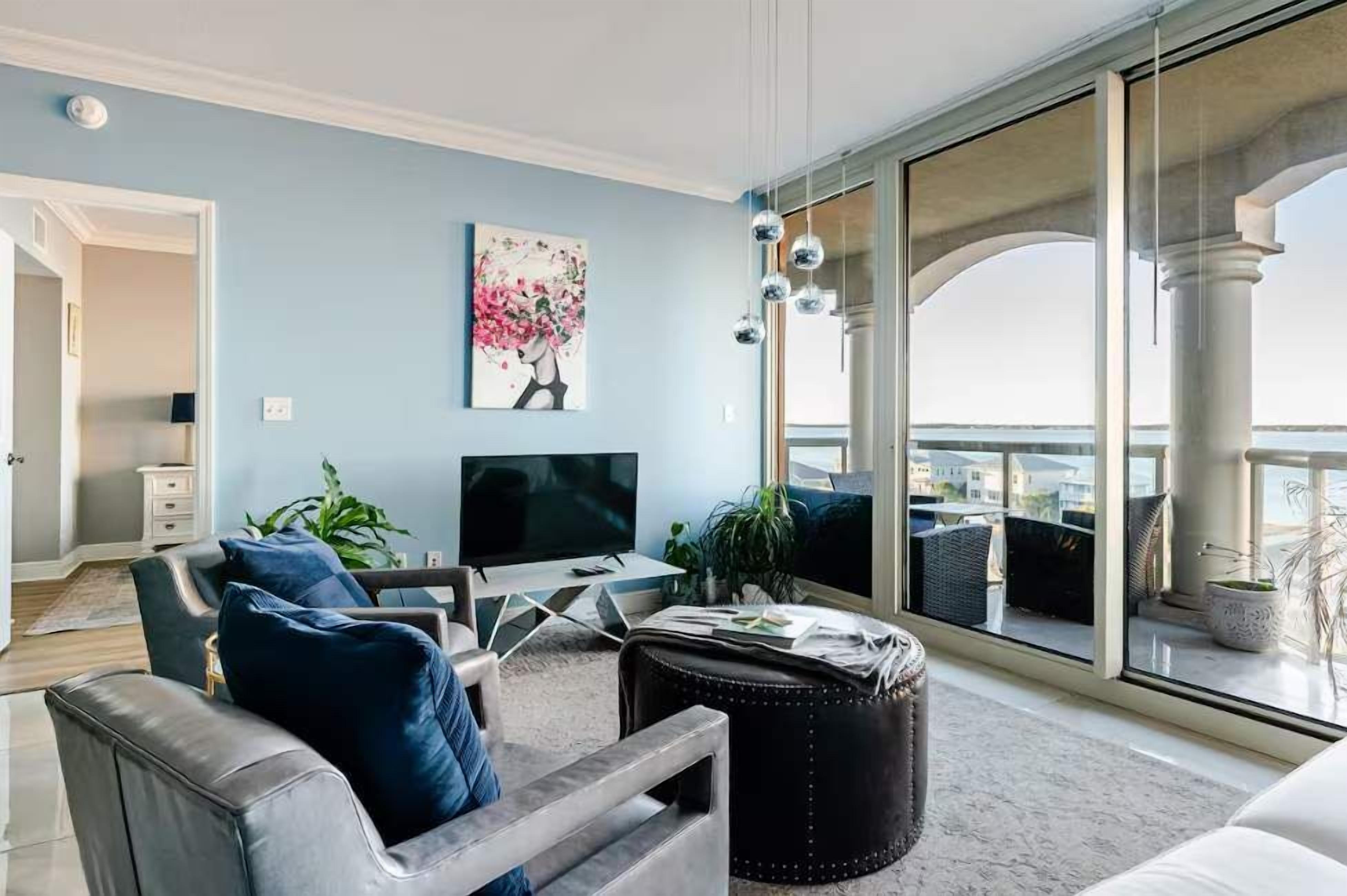 Una sala de estar con televisor, asientos, un cuadro y vistas al exterior.