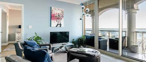 Una sala de estar con televisor, asientos, un cuadro y vistas al exterior.