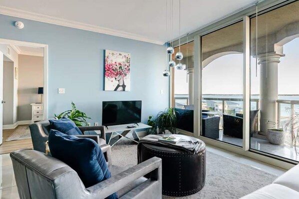 A living room with a TV, seating, a painting, and a view of the outside. - Ocean Front Condo w/ Pools, Sauna, Tennis & Gym (Escambia County)