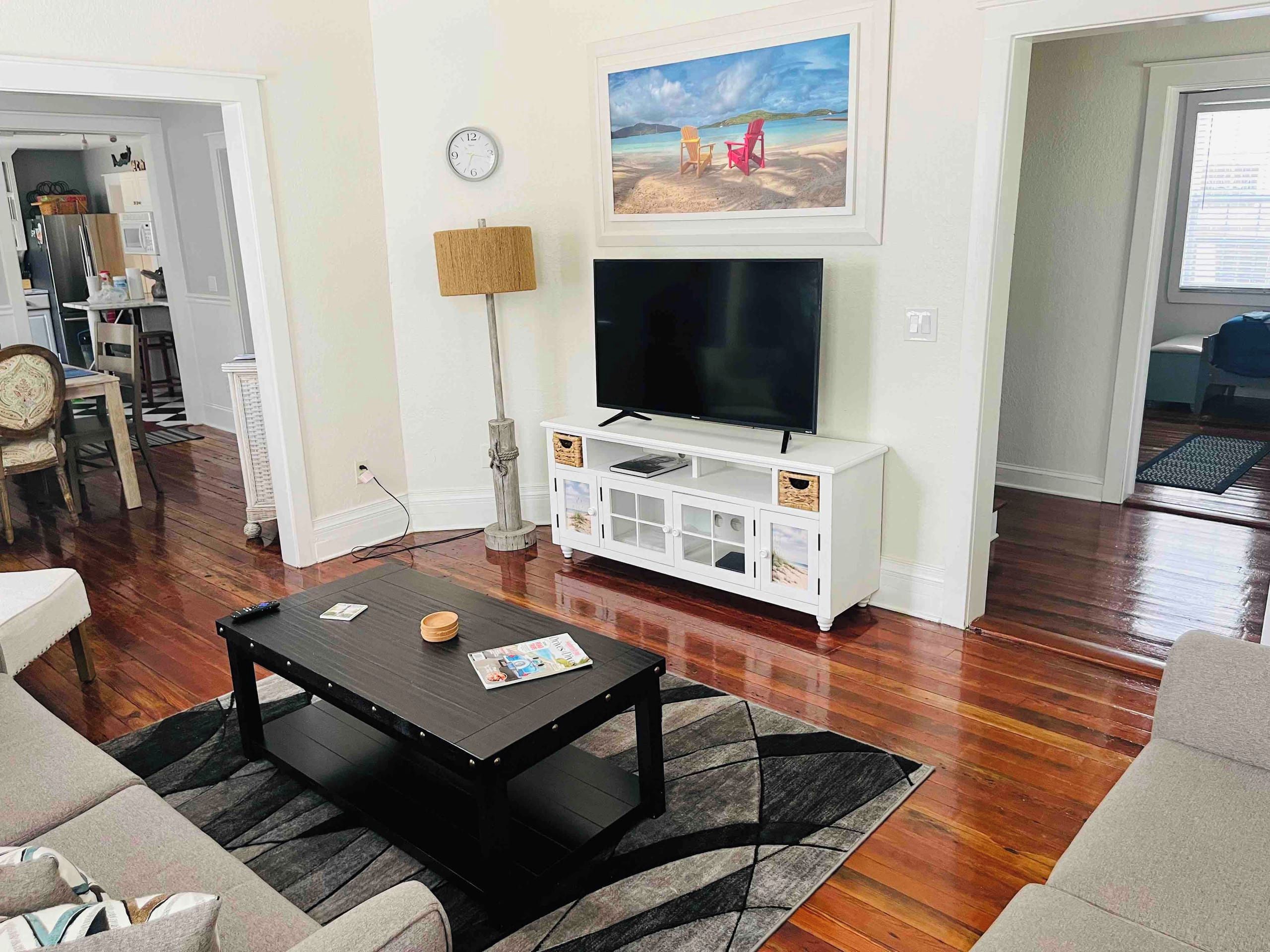 Una sala de estar con una televisión sobre un mueble blanco, una mesa de centro, un reloj y un cuadro con una playa.